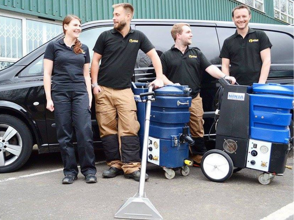 This is a photo of Rickmansworth Carpet Cleaning carpet cleaners (three men and one woman) standing in fromt of their black van, with two steam cleaning carpet machines next to them